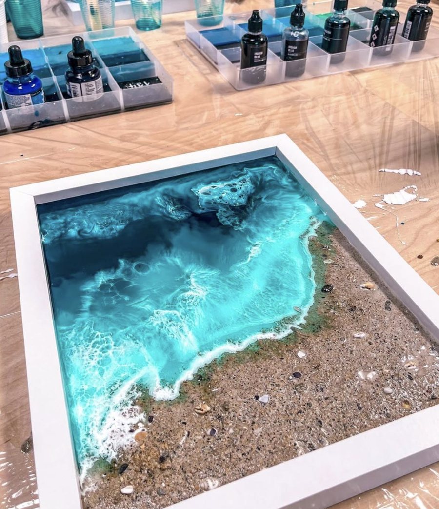 A white-framed resin ocean piece on the studio bench — turquoise water breaking onto real Cornish sand and shells.