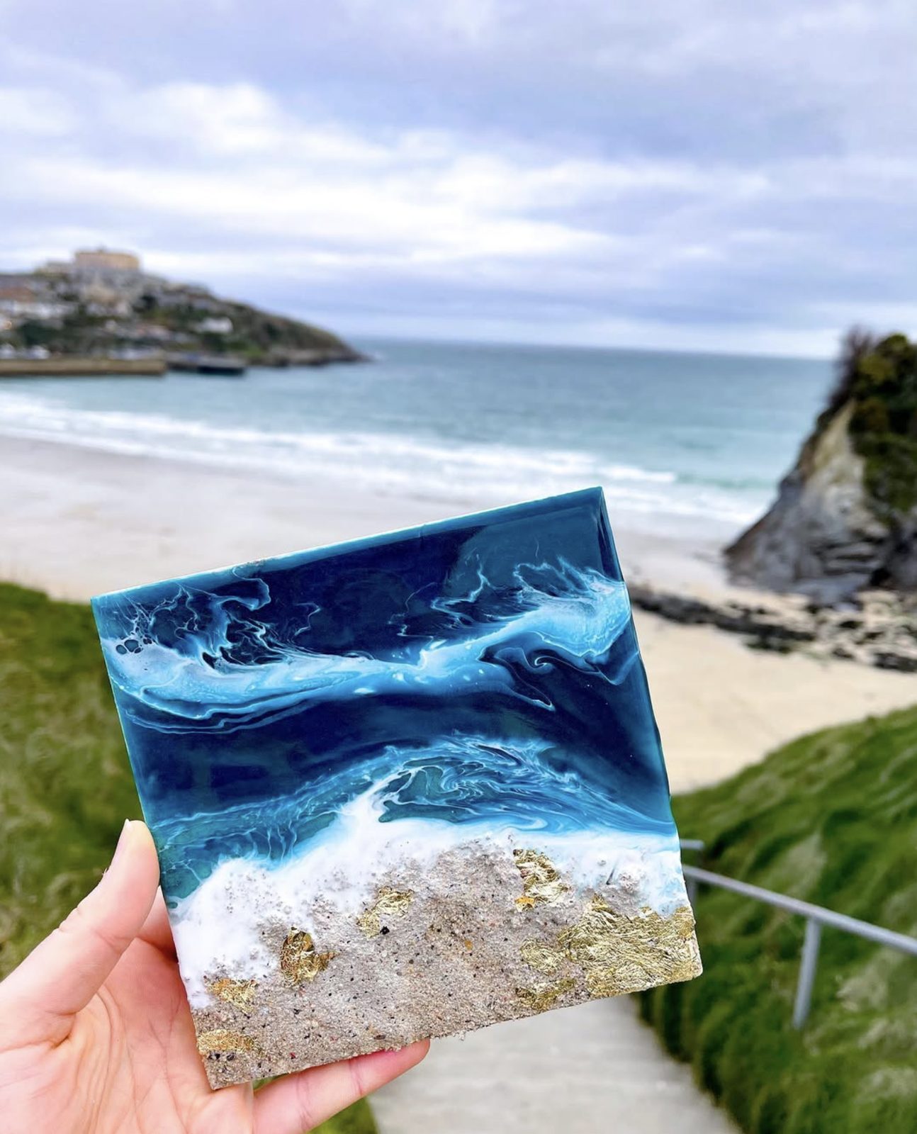 A finished resin piece held up above Towan beach — the coastline that inspired it behind.
